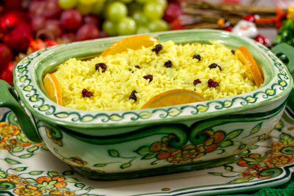 Ilustração vetorial plana em tons amarelo, laranja e verde de um prato cerâmico verde e amarelo com arroz amarelo guarnecido com rodelas de laranja e cranberries secas, sobre uma toalha decorada.